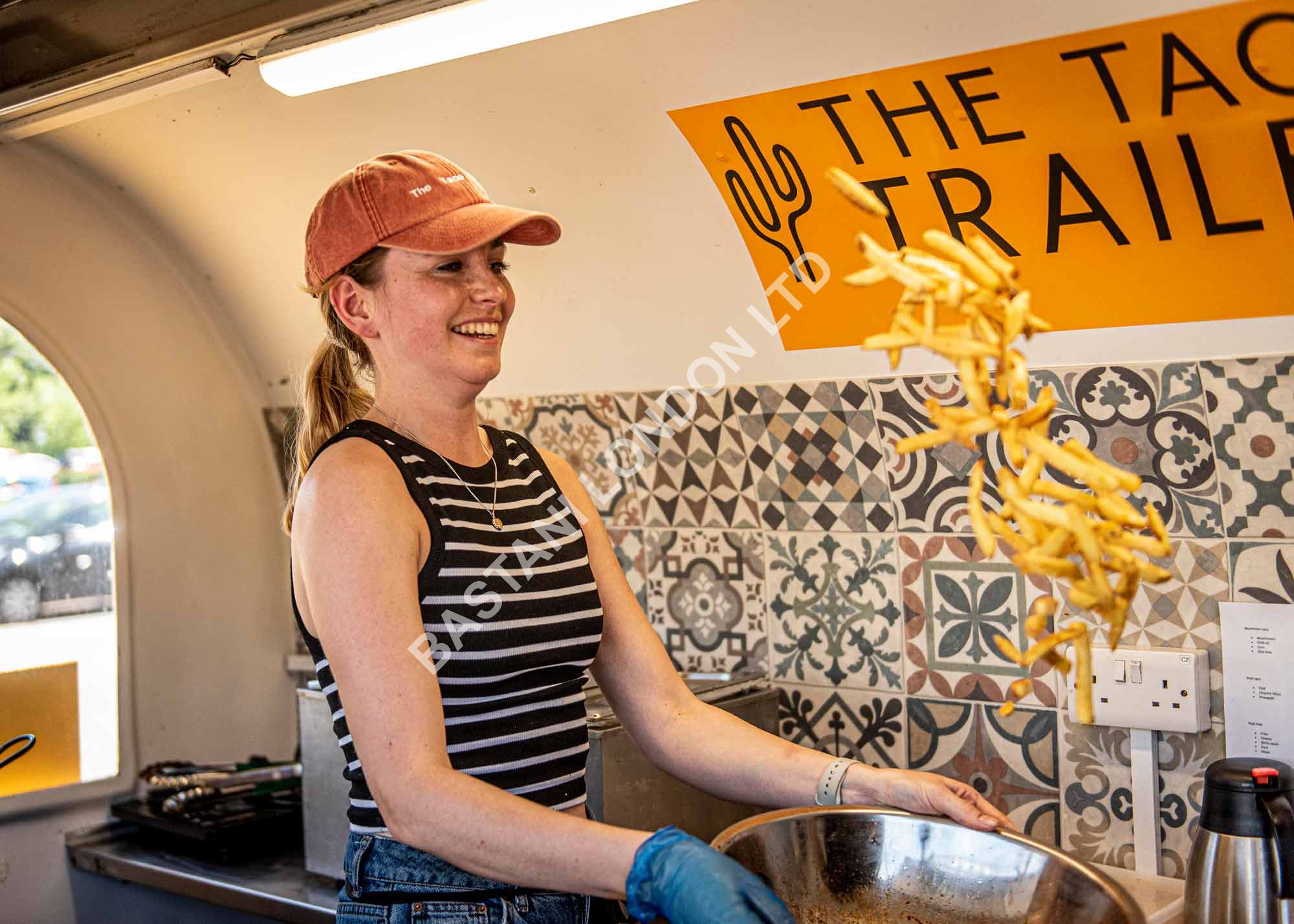 Smiling woman tossing fries at 'The BASTANI LONDON LTD' food stand